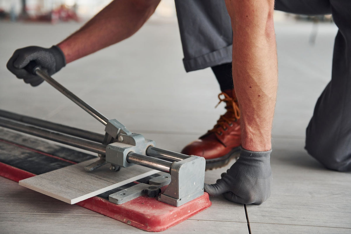 Worker using manual tile cutter to score light grey wood-grain tile, wearing work gloves and boots on tiled floor.