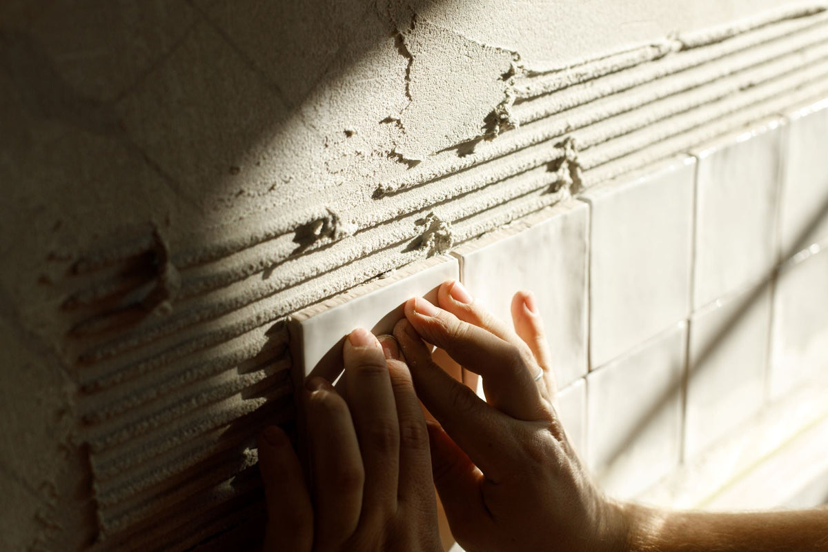 Tile installation in progress showing hands pressing white tile into grey adhesive with horizontal ridges on wall