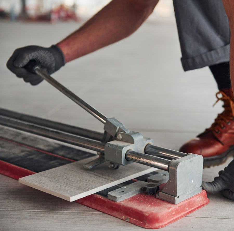 Worker using manual tile cutter to score light grey wood-grain tile, wearing work gloves and boots on tiled floor.