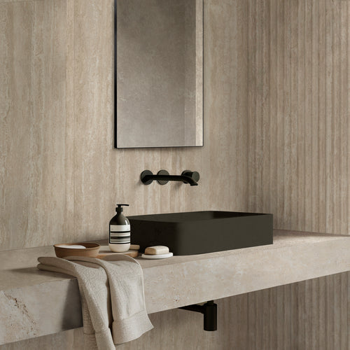 Slightly dim bathroom vignette with soap, towels, and a mirror beside Travertine Veincut Dorica 3D Beige Matte textured tile.