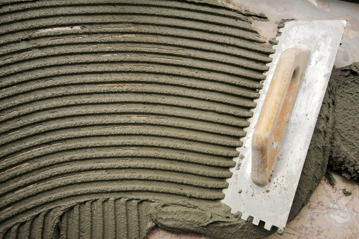 Metal notched trowel spreading grey tile adhesive in parallel ridges, demonstrating thinset and mortar application technique